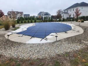 A covered in-ground pool with a blue safety cover, surrounded by rocks and landscaping, in a suburban backyard on a cloudy day.